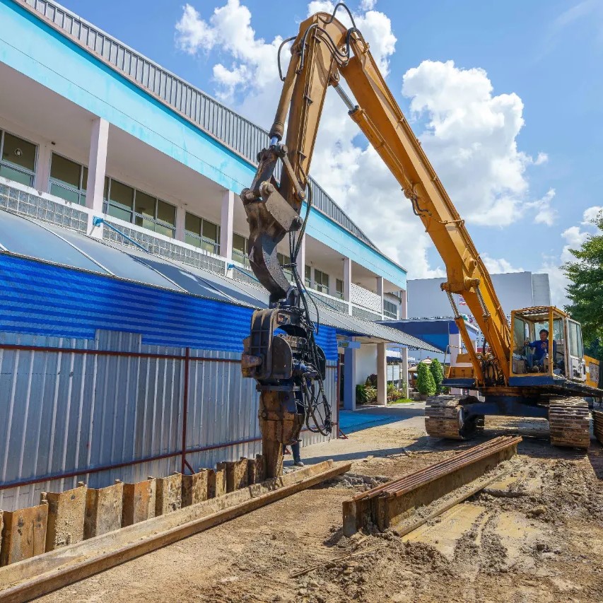 Steel Sheet Piling Services in Long Island - An Escavator Installing Sheet Piling Near a Commercial Building to Prevent Hydrostatic Pressuere